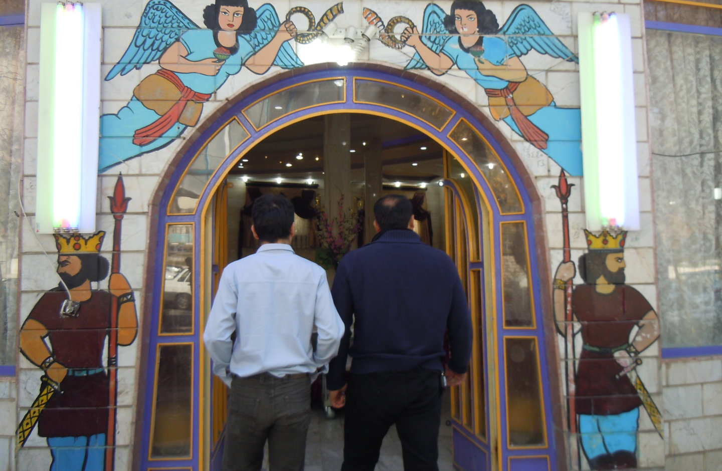 Two men from behind at the entrance to a restaurant in Tāq-e Bostān.